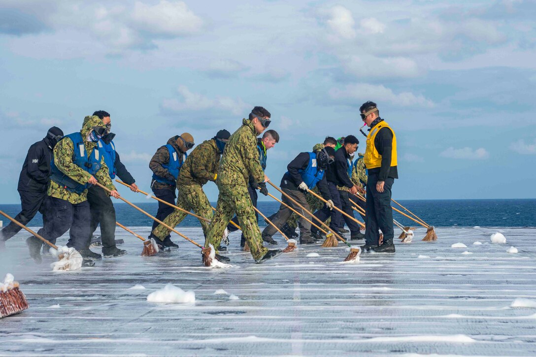 Sailors with brooms scrub the deck of a ship at sea.