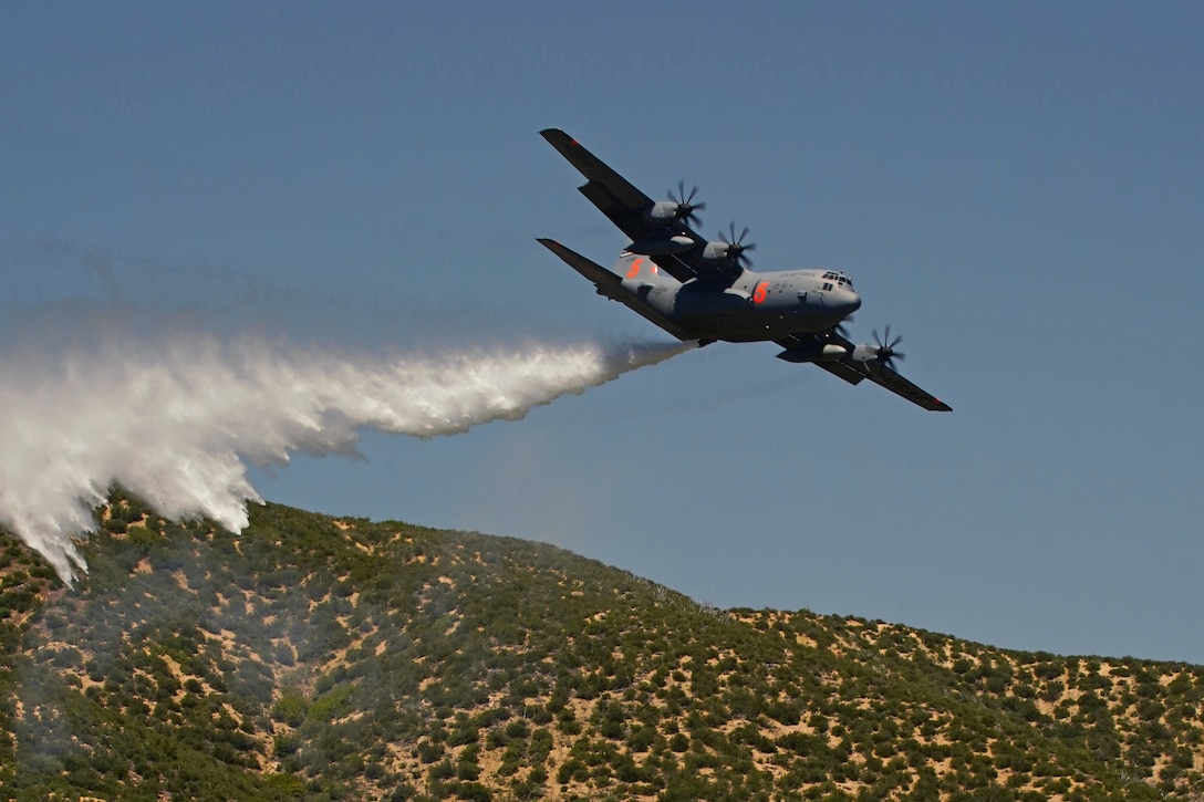 A military aircraft drops water over a forested area during training.