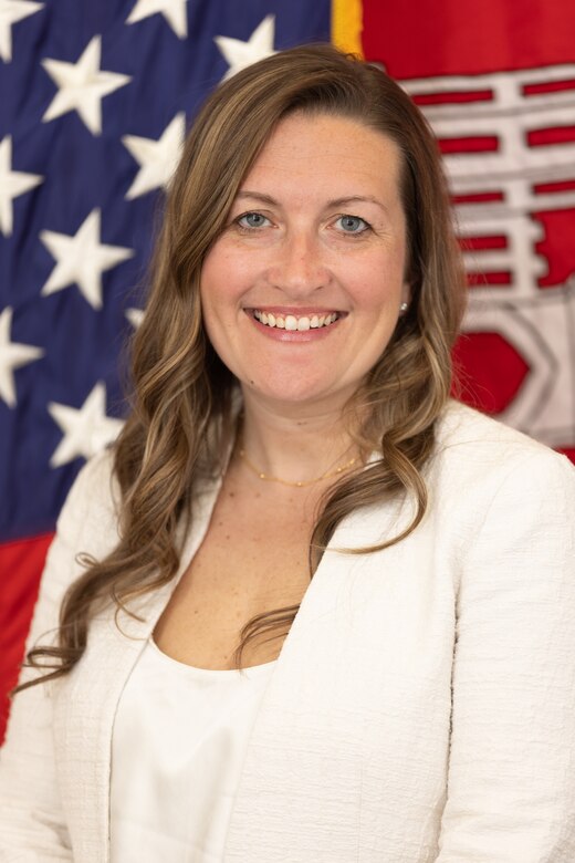 A woman, Ms. Liz Walker, in a white jacket and white shirt smiles in front of the U.S. flag and the red and gold castle flag of the U.S. Army Corps of Engineers.
