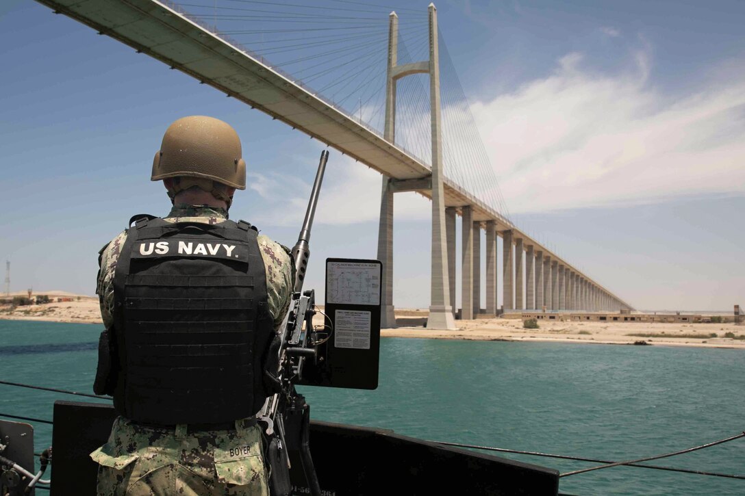 A sailor stands next to a weapon aboard a ship while transiting a body of water under a bridge.