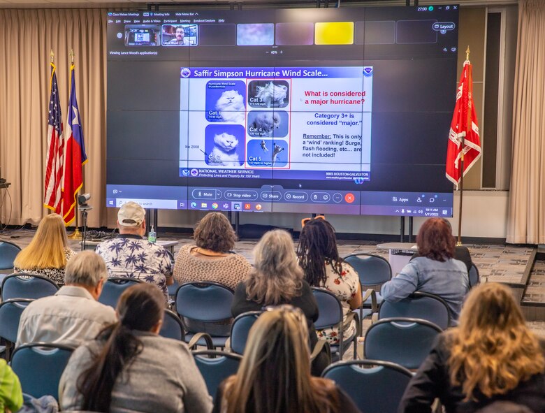 a group of people in the foreground are seated looking at a large screen with a PowerPoint slide with different funny photos of cats use to describe categories of hurricanes.