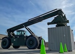 A large piece of green equipment with an extendable boom prepares to lift a container that is used to ship items on the back of semitrucks. The operation is taking place in a parking lot.