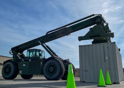 A large piece of green equipment with an extendable boom prepares to lift a container that is used to ship items on the back of semitrucks. The operation is taking place in a parking lot.