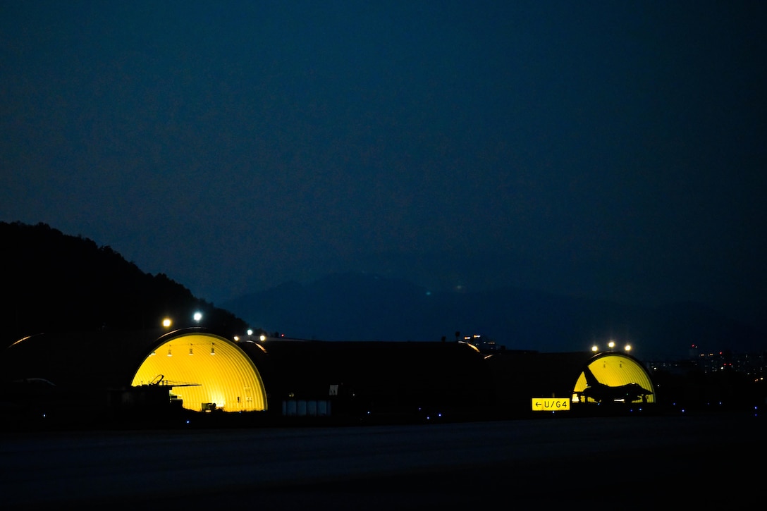 A fighter jet sits outside two hangars lit up against a dark night.