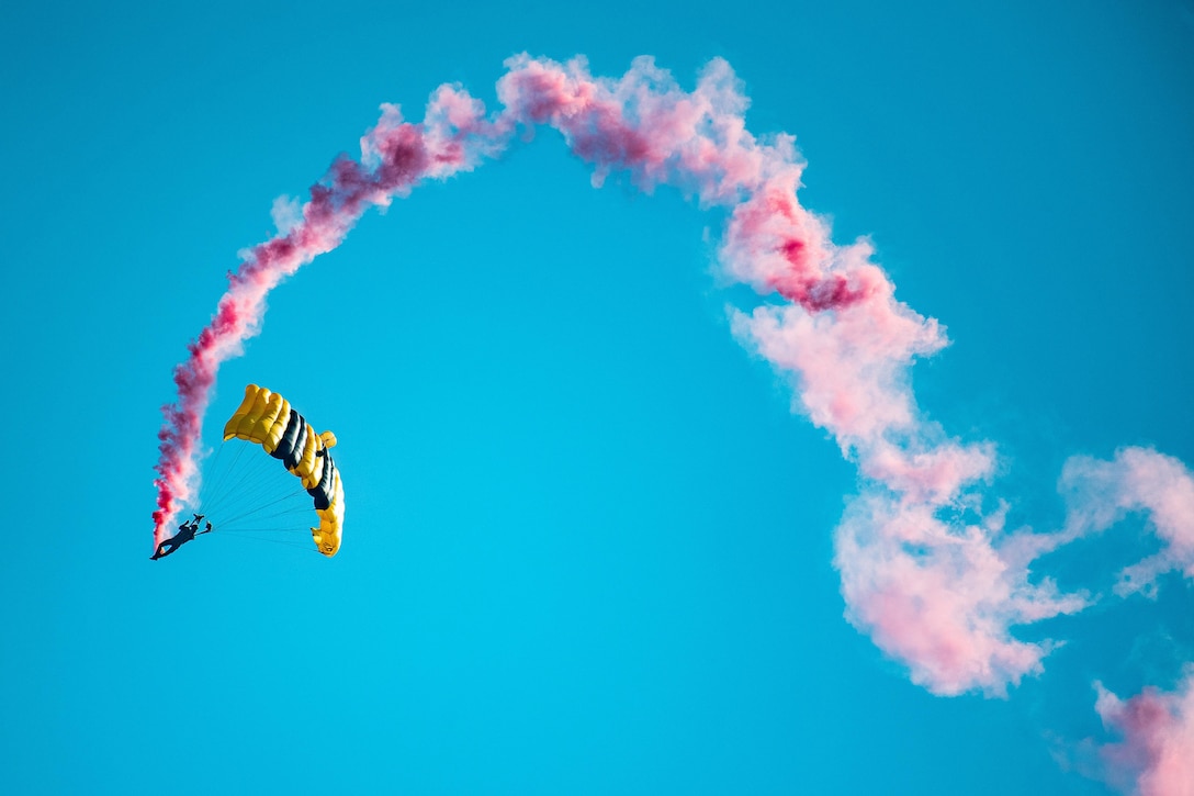 A soldier performs a parachute drop trailing pink smoke against a blue sky.