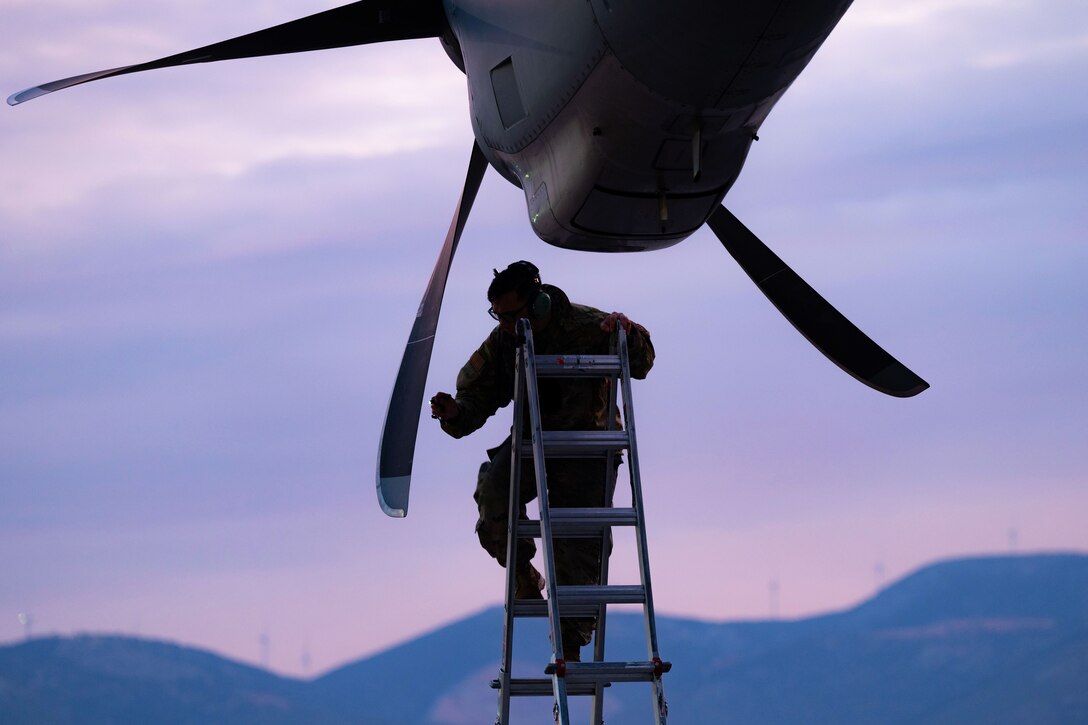 An airman stands on a ladder and inspects an aircraft propeller.