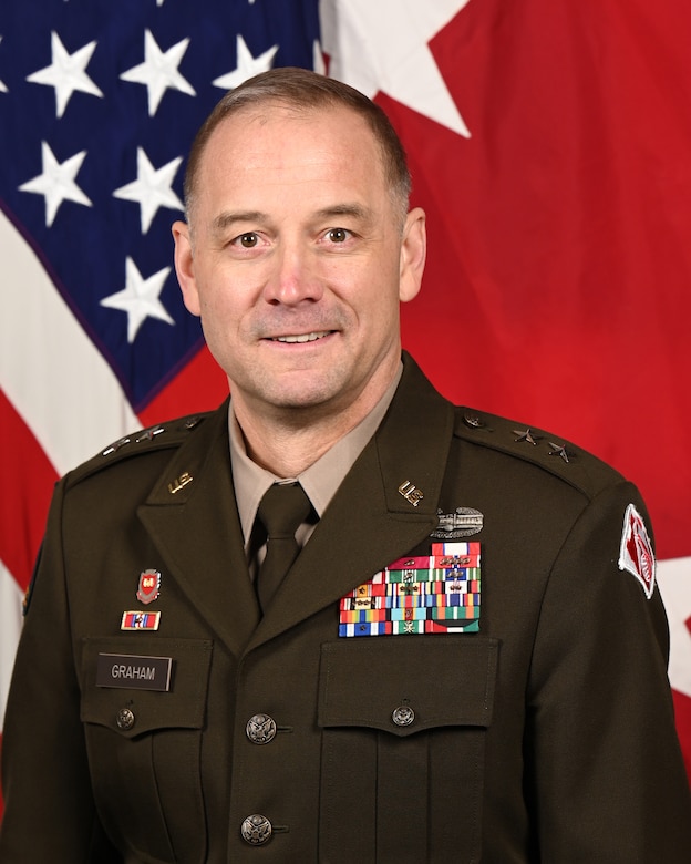 A man, Major General William (Butch) H. Graham, in a dark green Army uniform poses in front of the U.S. Flag and the a red castle flag representing the U.S. Army Corps of Engineers.