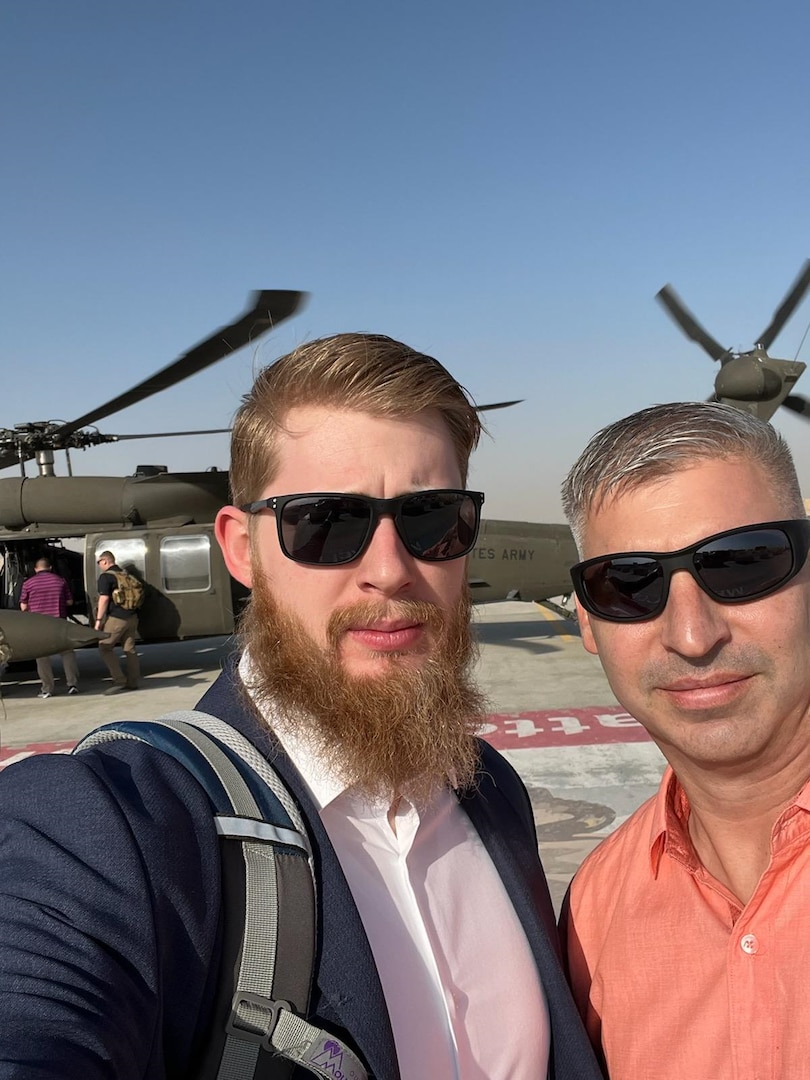 Two smiling men with sunglasses stand near helicopters. One wears a melon Polo shirt and the other a white dress shirt and a blue suit coat. One has red hair and a beard ad the other has graying hair cut short. Both are light skinned.