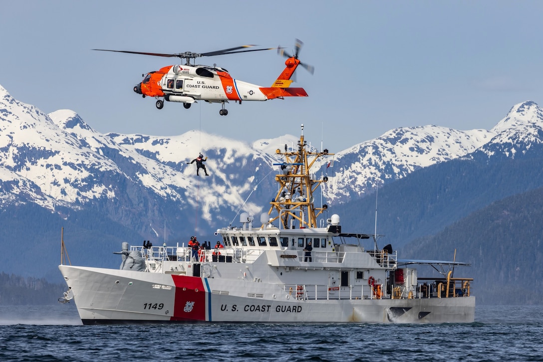 A rescuer hangs by a line under a helicopter hovering above a cutter in water with snowy peaks in the background.