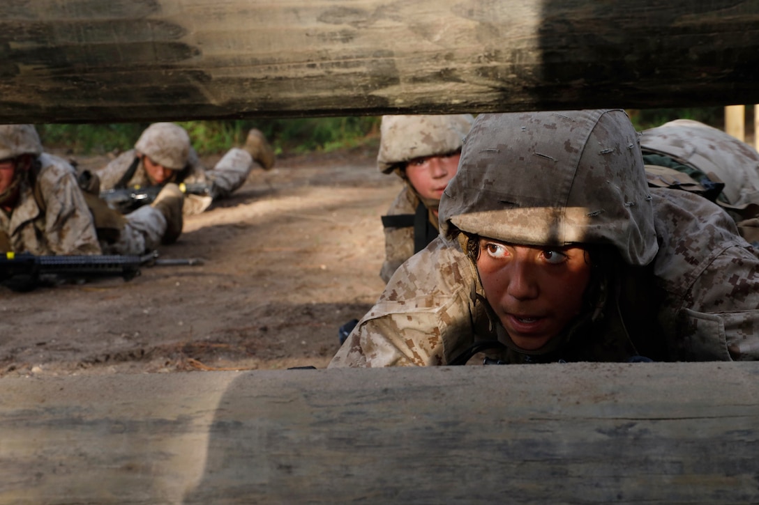 Marine Corps recruits crawl on the ground with weapons while looking through an obstacle course.
