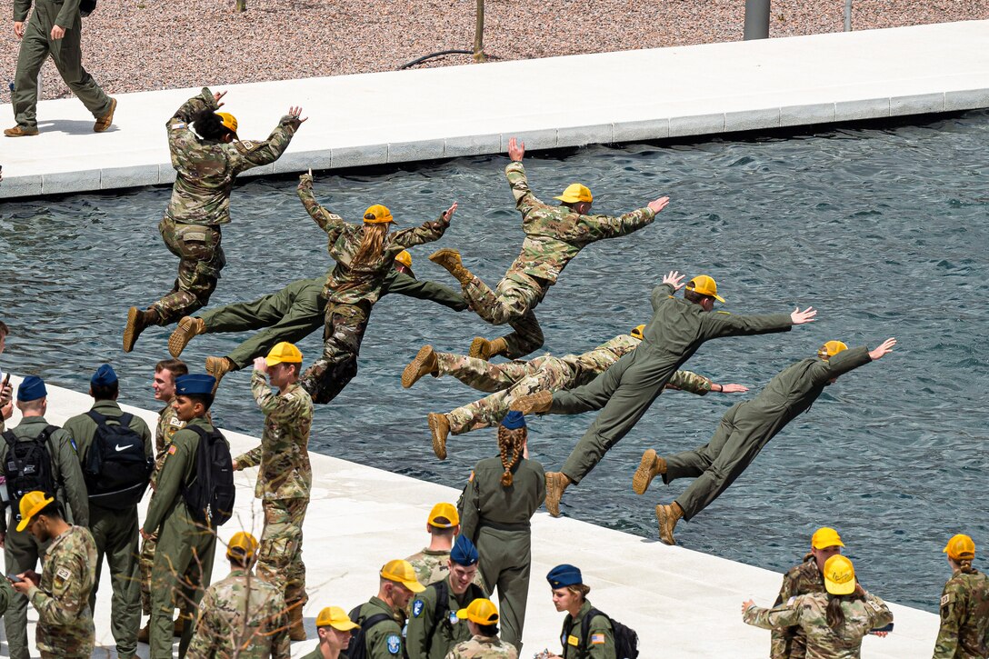 Air Force cadets wearing yellow hats jump into a fountain as fellow cadets stand in the background.