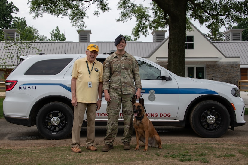 A man in uniform stands with another man and a dog in front of a police vehicle.