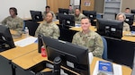 Male and female soldiers sit at desks in front of computers in a classroom.