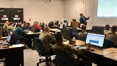 Soldiers and civilians sit in a classroom facing a projector screen where a teacher stands