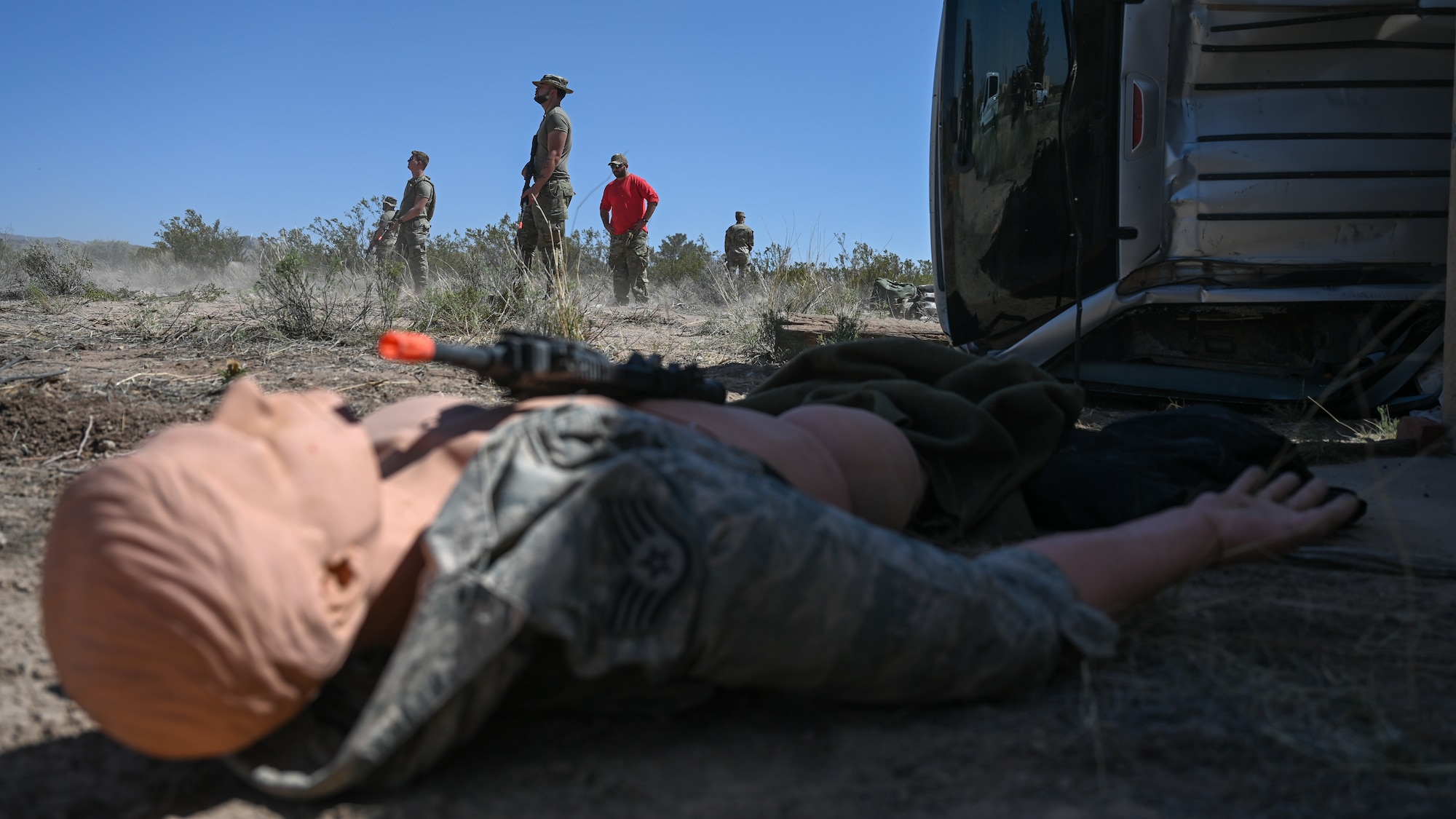 A mannequin lays on the ground while people in military uniforms stand in the distance.