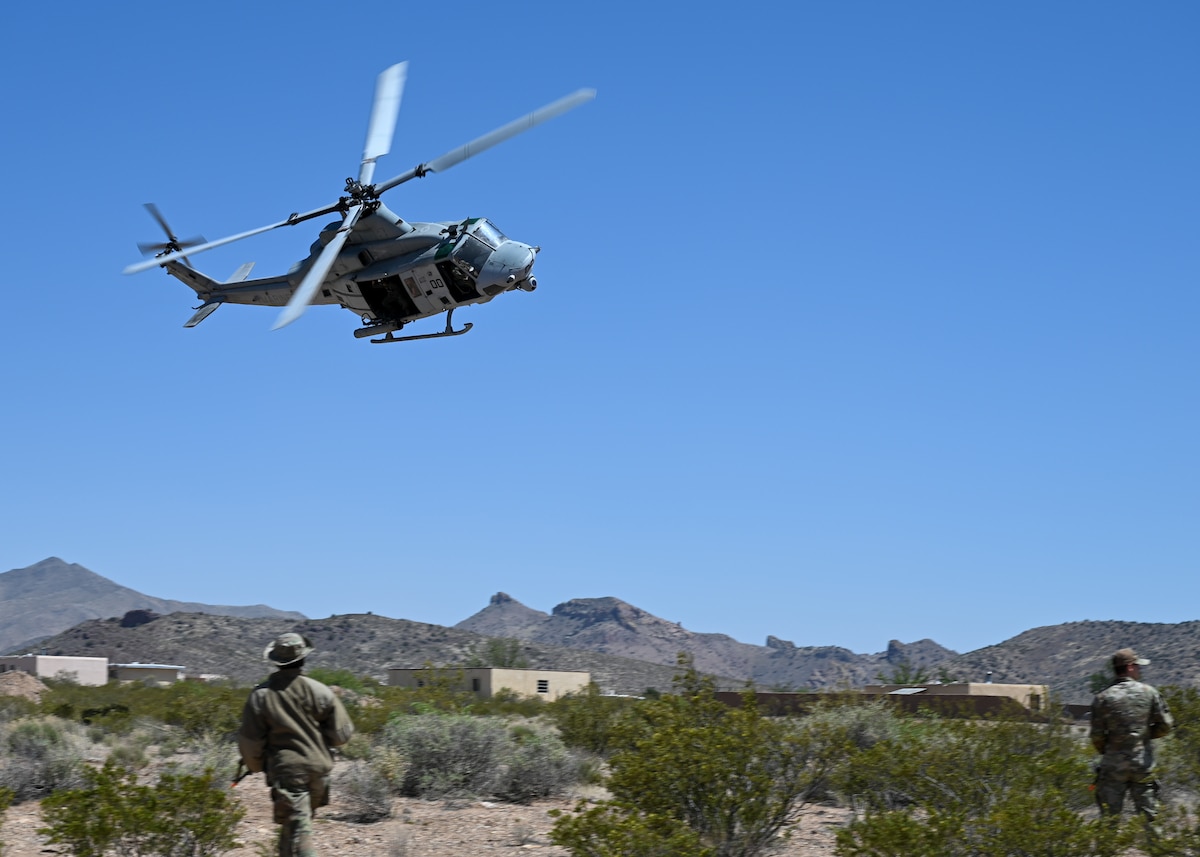 People in military uniforms stand in an open area as a helicopter flies close by.