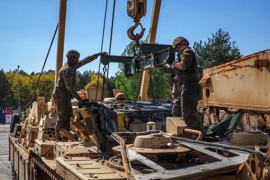 Two soldiers pull an engine from a tank during maintenance while another soldier watches.