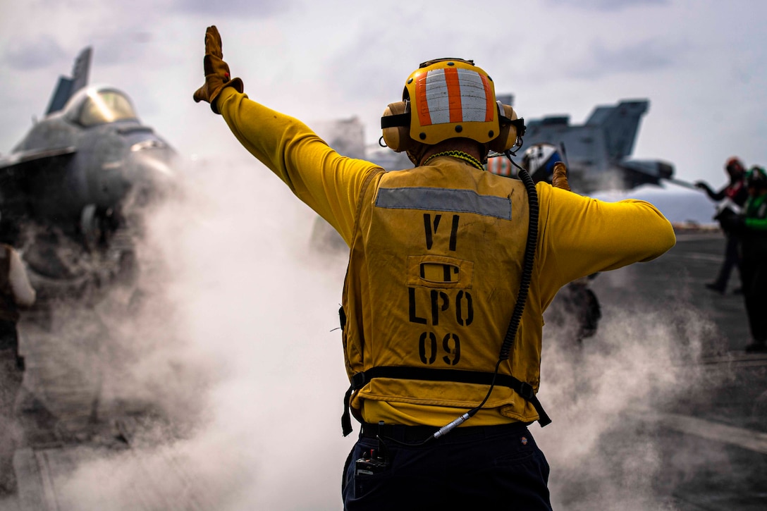 A sailor signals toward an aircraft as smoke surrounds.
