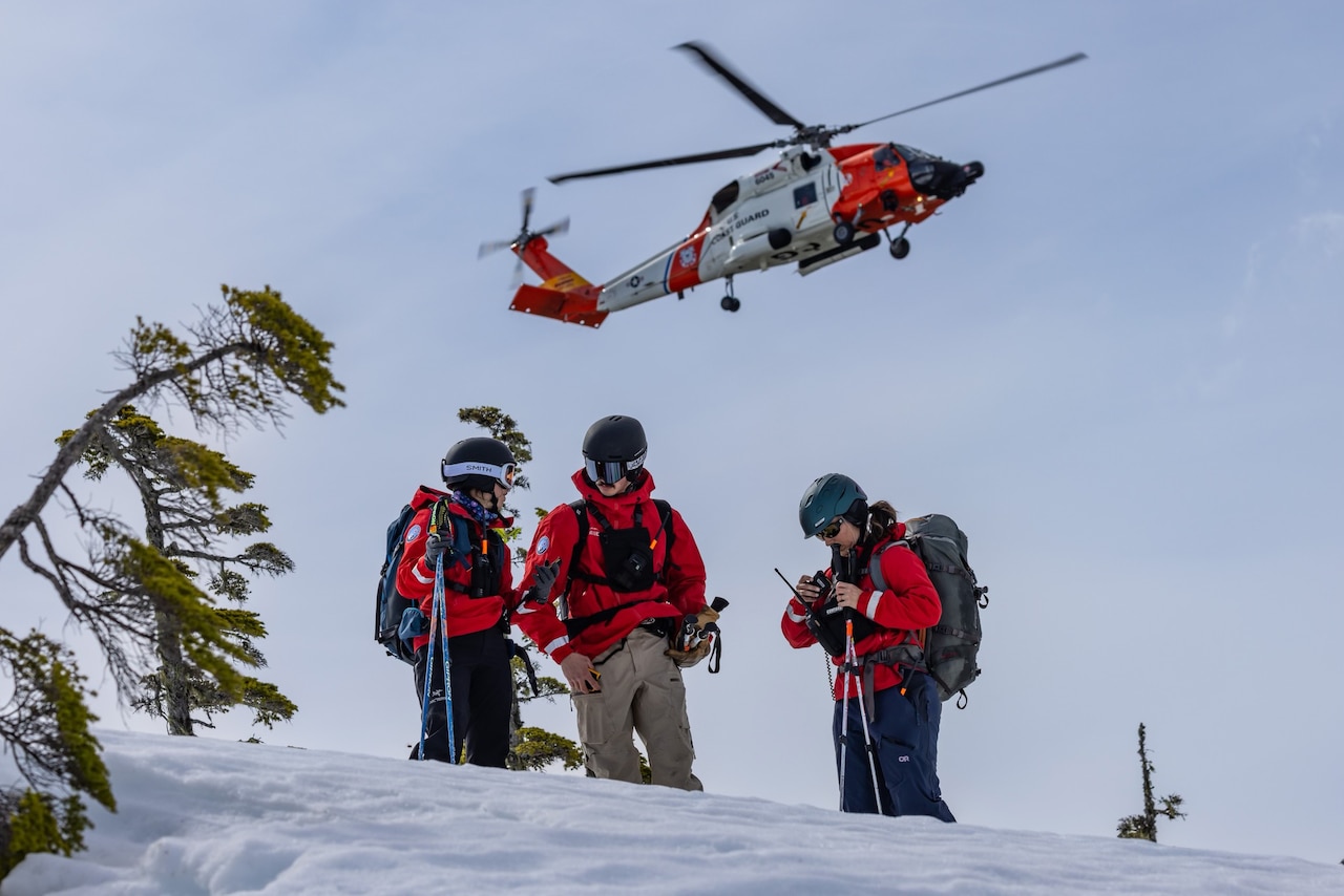 Three people in red jackets with ski poles stand and confer in the snow as a helicopter flies above.