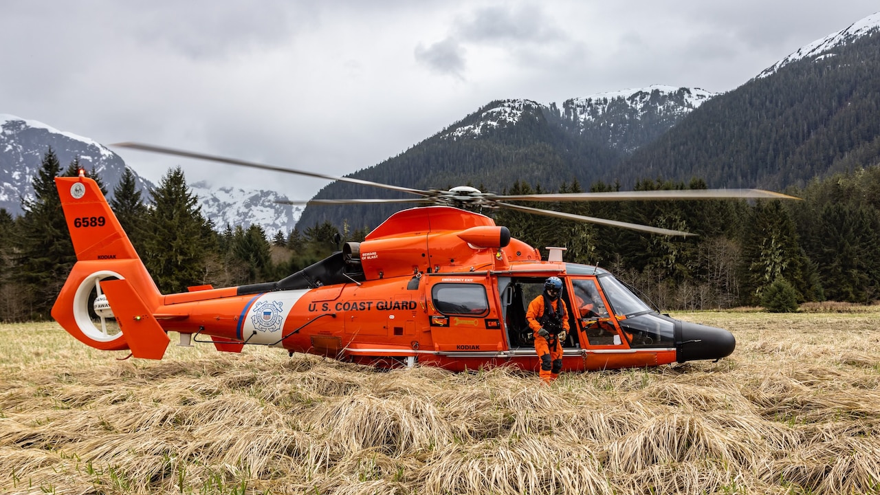 A crew member stands outside an orange Coast Guard helicopter parked in a field.