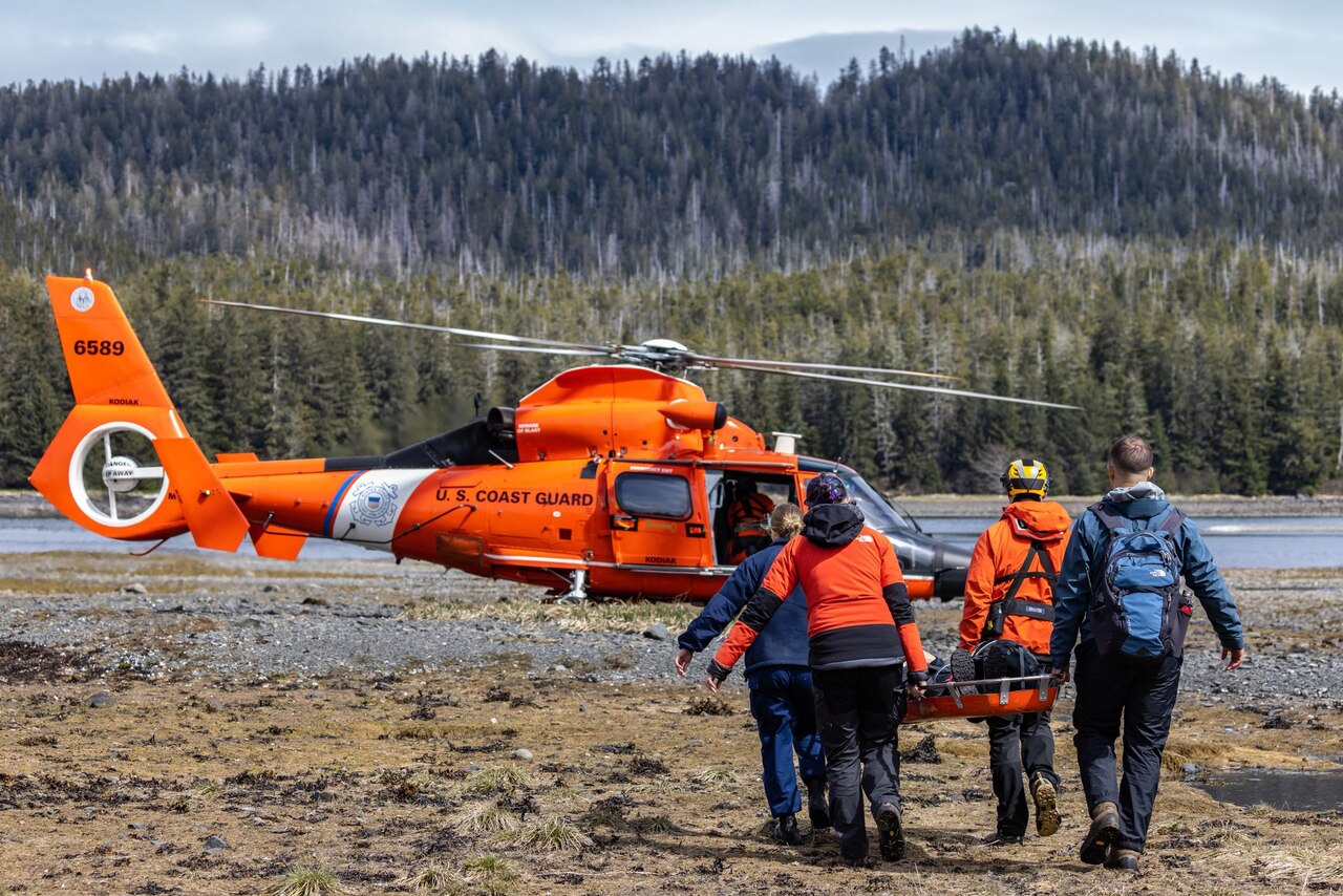 Four people carry a litter toward the open door of a parked Coast Guard helicopter.