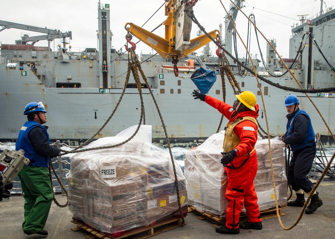 Sailors aboard a ship remove rope from pallets of cargo as a ship sails in the background.