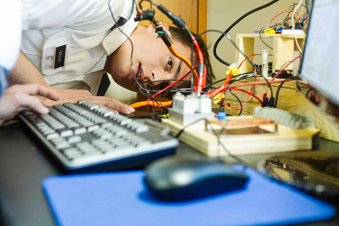 A U.S. Military Academy cadet rests their head on a table to look at wires attached to a gadget as a person types on a keyboard.