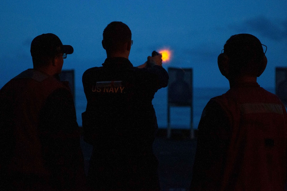 A sailor fires a weapon aboard a ship at a target as two sailors watch illuminated by orange light.