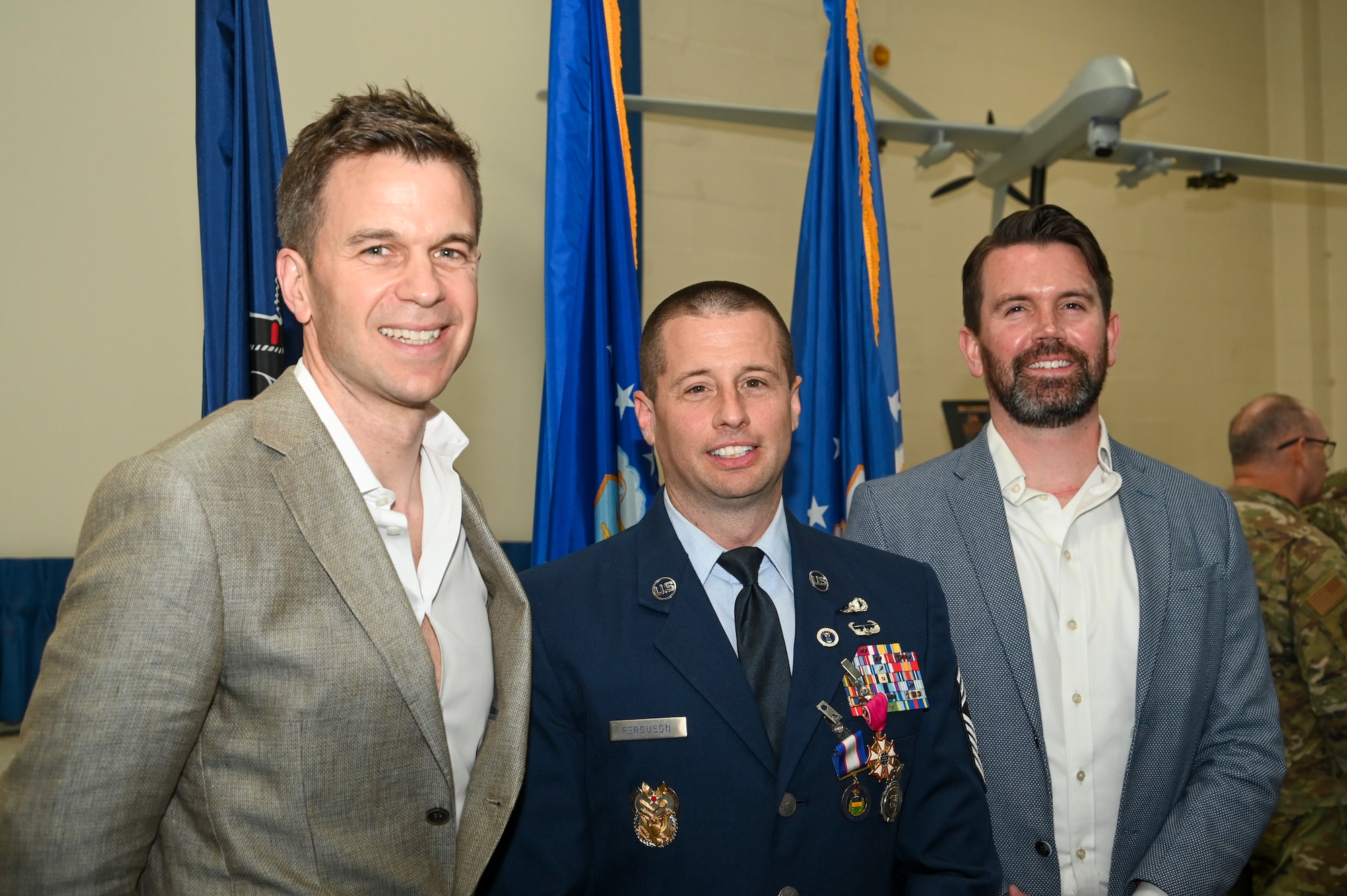 Three men pose for a photo. The man in the center is wearing Air Force Dress Blues.