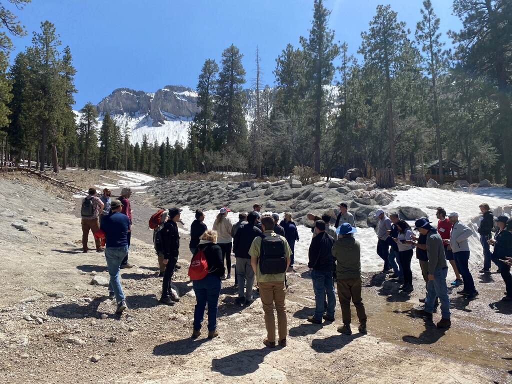 Tim Fairbank, U.S. Army Corps of Engineers (USACE), Los Angeles District, describes the design of a berm and diversion channel constructed to protect residents of the Rainbow Canyon subdivision (near background) following a series of debris flows associated with the 2013 Carpenter One fire in the Spring Mountains, NV. Portions of the burn scar are visible on the mountainside in the background. The photo includes the attendees of the first USACE CWMS (Corps Water Management System) wildfire Workshop held in Las Vegas, NV on April 24-27, 2023.