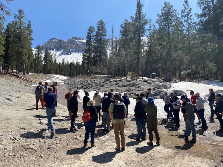 Tim Fairbank, U.S. Army Corps of Engineers (USACE), Los Angeles District, describes the design of a berm and diversion channel constructed to protect residents of the Rainbow Canyon subdivision (near background) following a series of debris flows associated with the 2013 Carpenter One fire in the Spring Mountains, NV. Portions of the burn scar are visible on the mountainside in the background. The photo includes the attendees of the first USACE CWMS (Corps Water Management System) wildfire Workshop held in Las Vegas, NV on April 24-27, 2023.