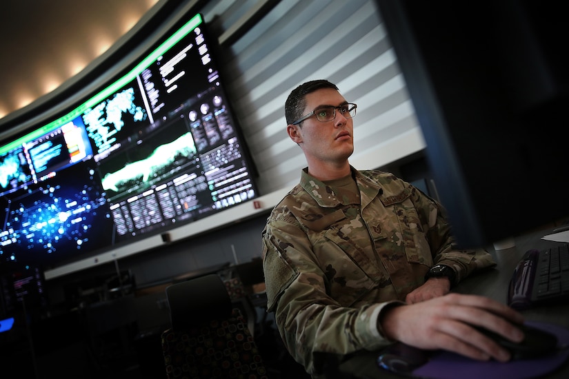 A man in uniform is seated behind a computer with a large watch floor display in the background.