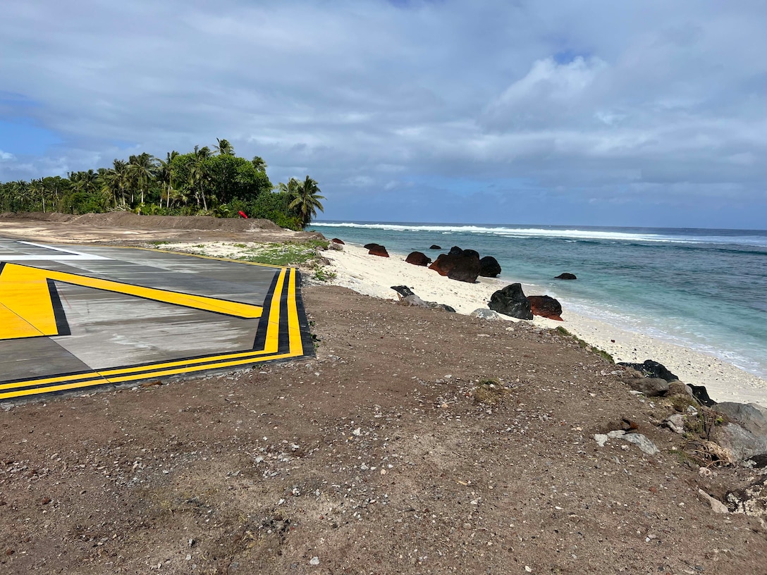 In the distance is a patch of green palm trees. Sky is covered in low white clouds. In the foreground yellow and black lines on concrete outline the perimeter of the edge of an airport runway that sits at a shoreline. To the right is a white sand beach and the blue ocean with no waves.