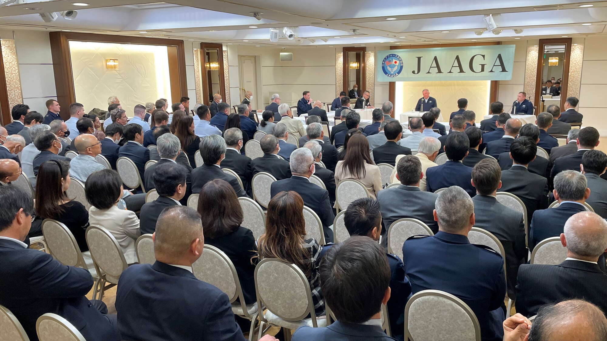 A crowded room of Japanese people watch two Americans speaking