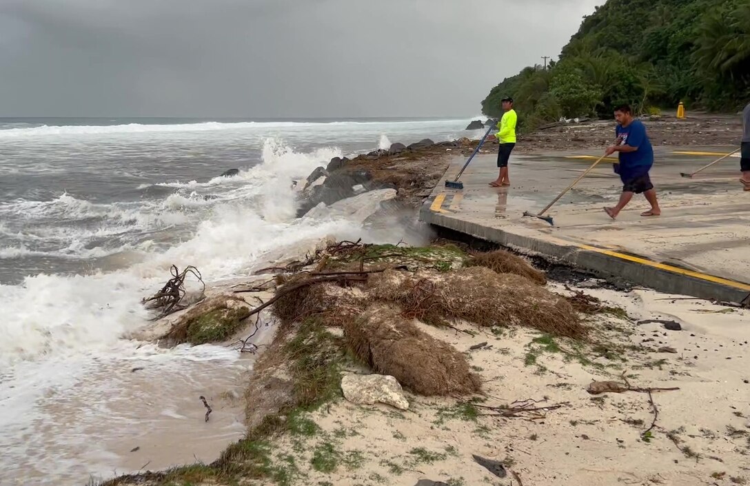 Two men facing the ocean push water off a concrete slab with brooms. Waves crash onto a concrete rocky shoreline.