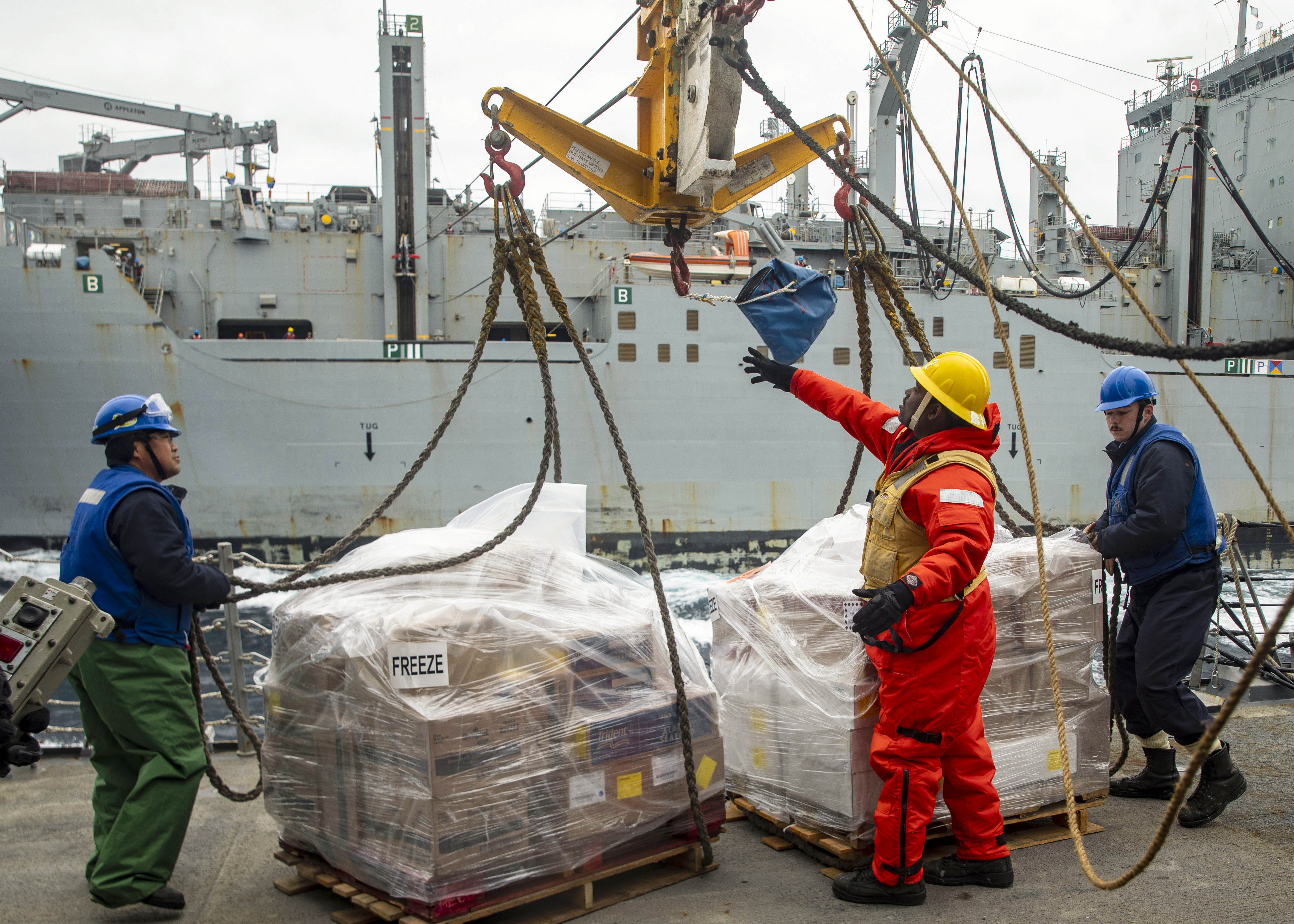 Sailors aboard a ship remove rope from pallets of cargo as a ship sails in the background.