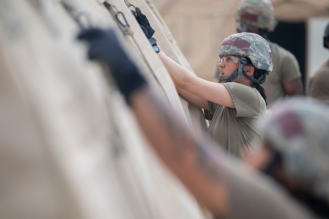 Several airmen, seen from close range, attach hooks to the side of a tent.