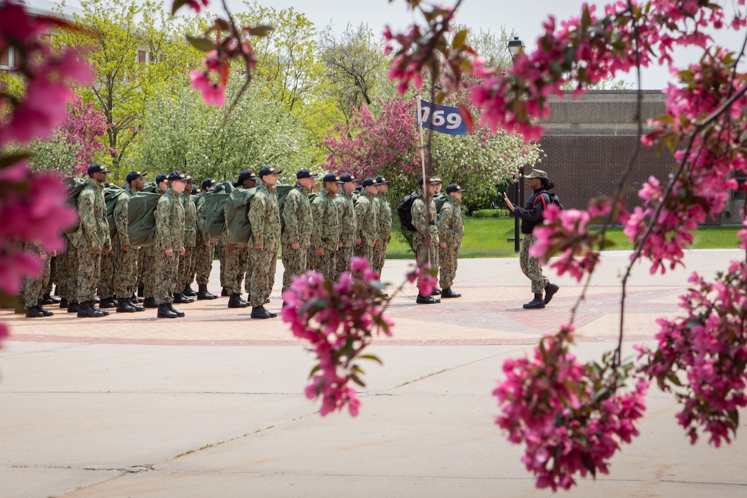 Navy recruits stand in formation framed by flowers on a tree.