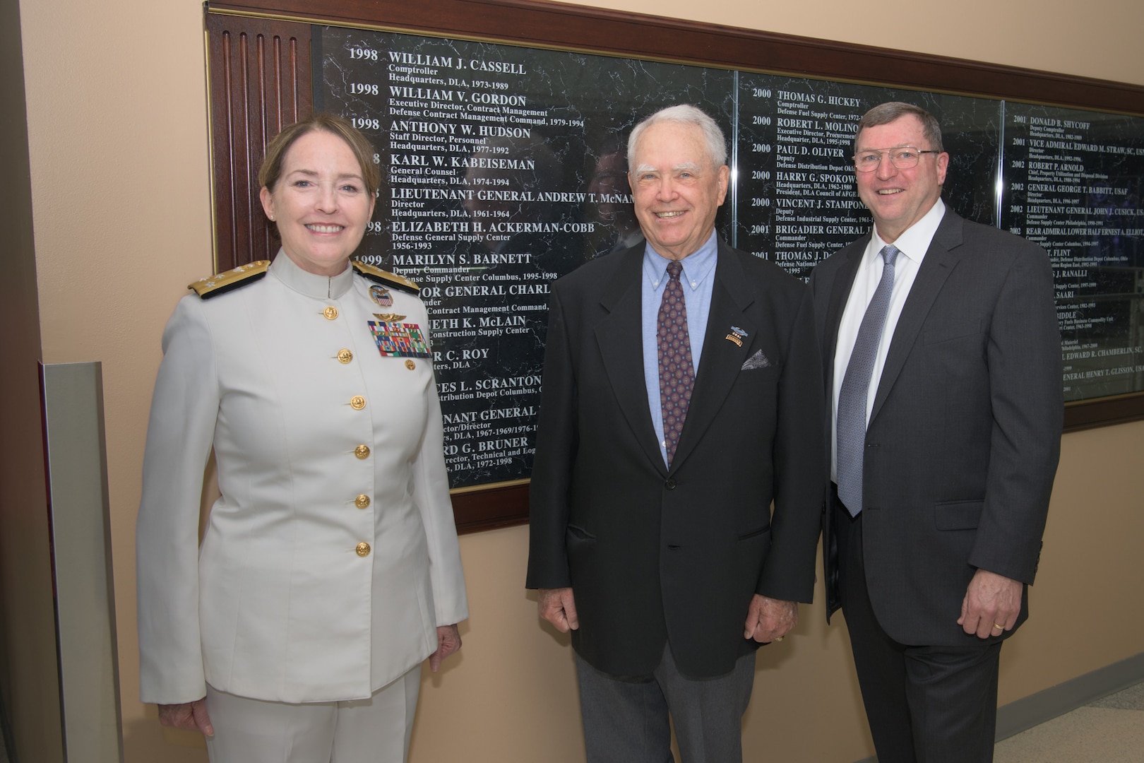 Three people pose in front of the Hall of Fame wall at the Defense Logistics Agency headquarters in Fort Belvoir, Virginia.