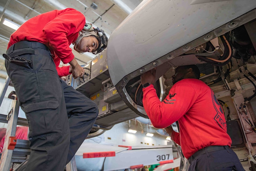 Two sailors work on a military aircraft inside a ship’s hangar bay.