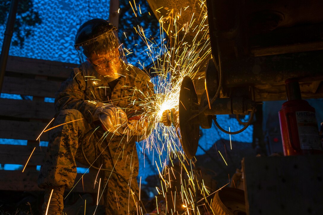Sparks fly as a Marine wearing protective gear kneels to work on a vehicle in the dark.