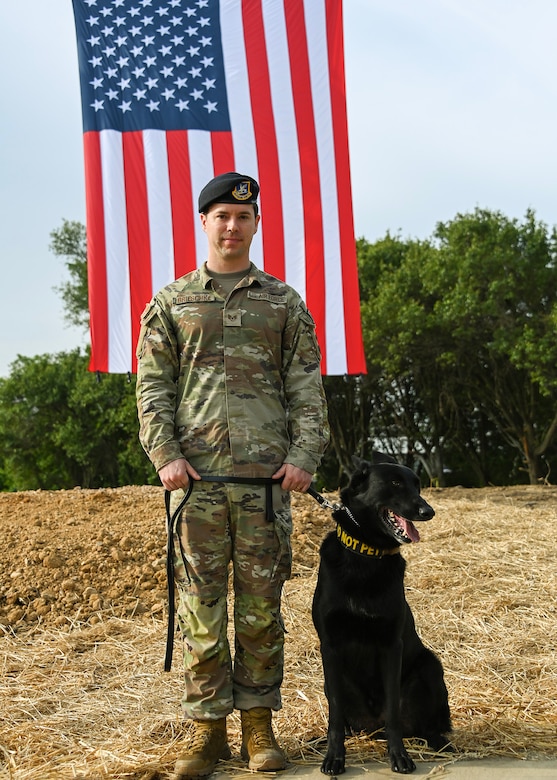 A man in military uniform stands next to a dog in front of a U.S. flag.