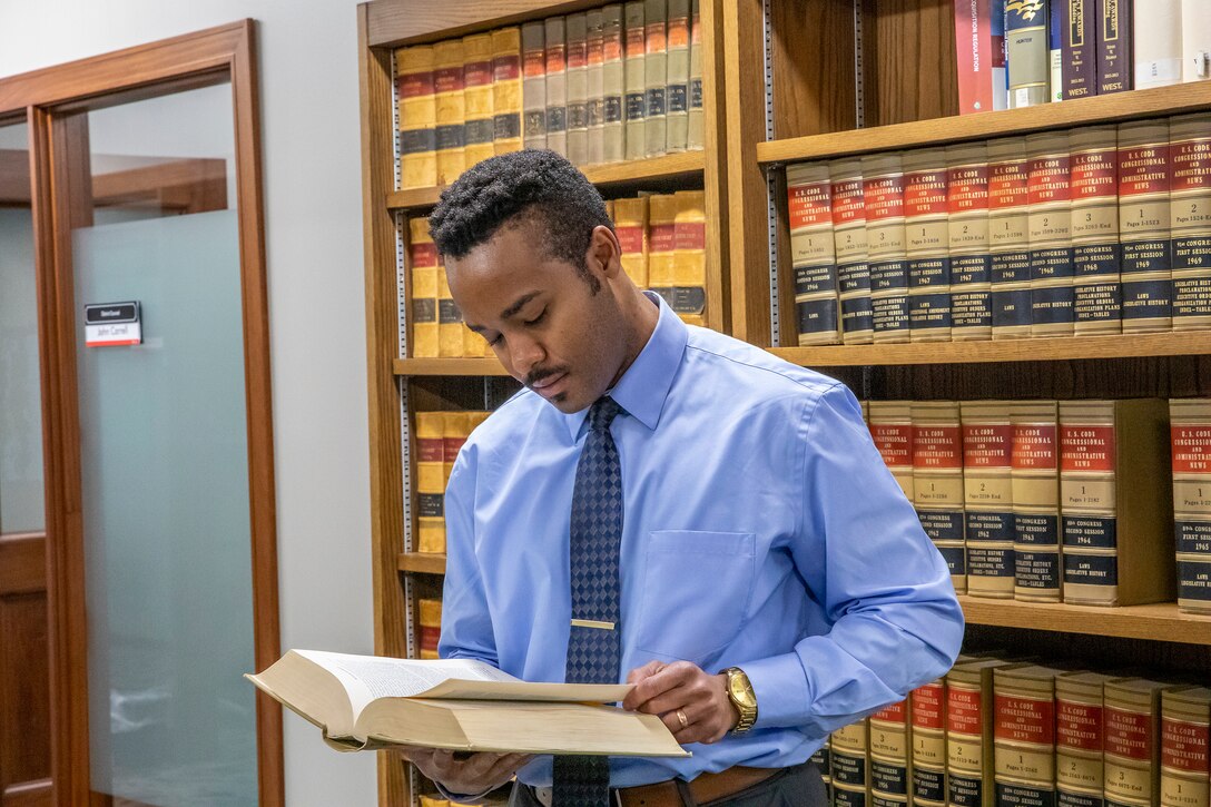 A man, Alex Webb, in a blue shirt and tie, reviews a legal book in front front of brown shelving with red, gold and black books inside of an office.
