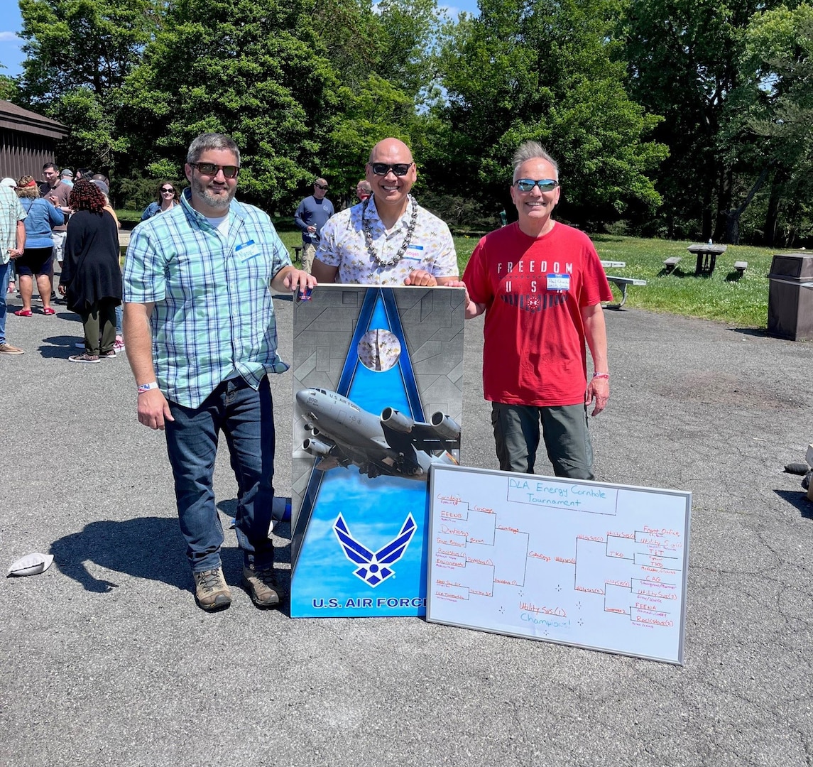 three gentlemen stand together with a cornhole board