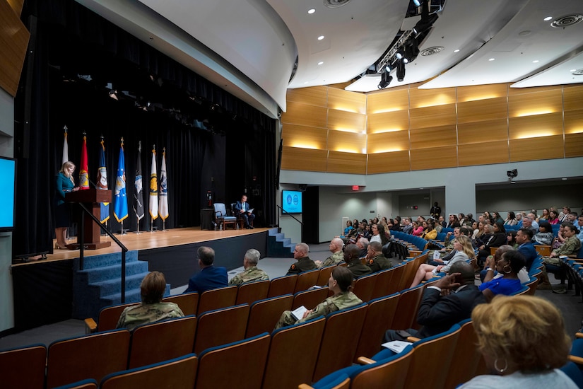 A woman speaks from a stage in an auditorium.