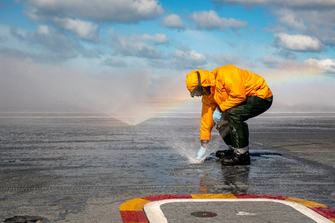 A sailor wearing rain gear unclogs an active sprinkler aboard a ship as sprinklers run in the background near a rainbow.