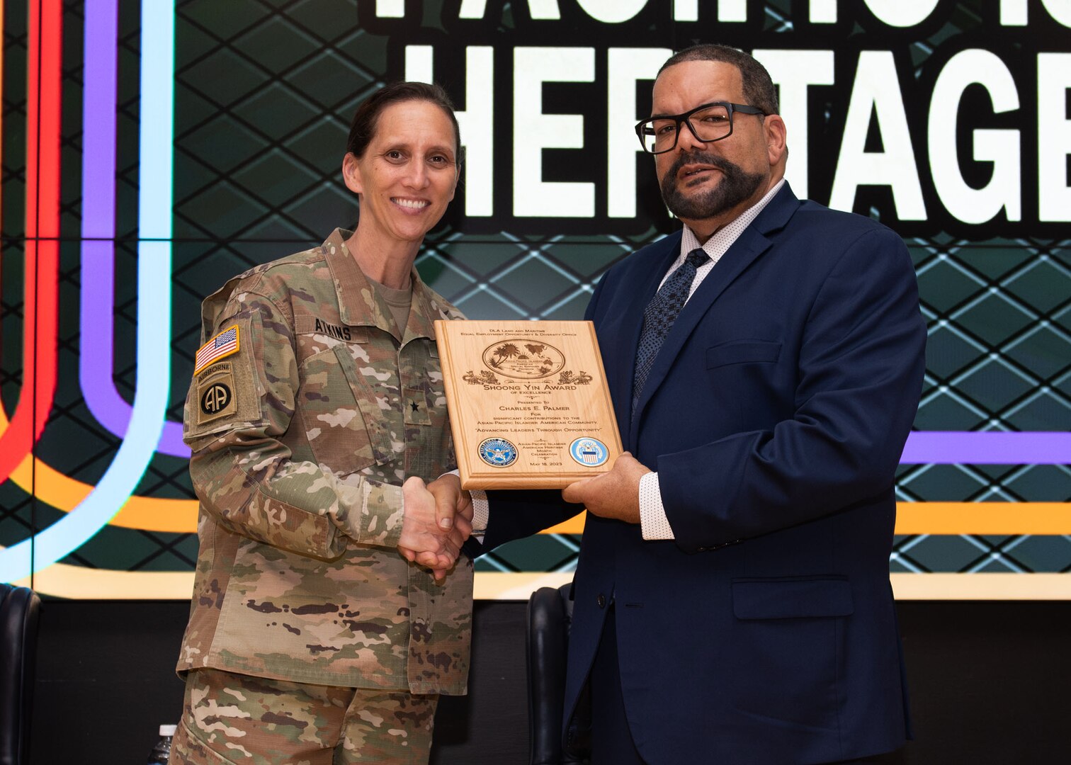 A light skinned dark haired woman in a camouflaged military uniform shakes hands with a darker skinned man with dark hair and a beard in a suit with glasses.