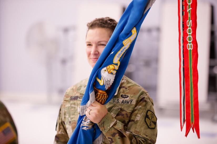 Lt. Col. Tenay Benes, a female Soldier, smiles as she holds the battalion colors in her hands. The colors are blue, with an eagle visible and multi-colored streamers hang off of the top of the staff.