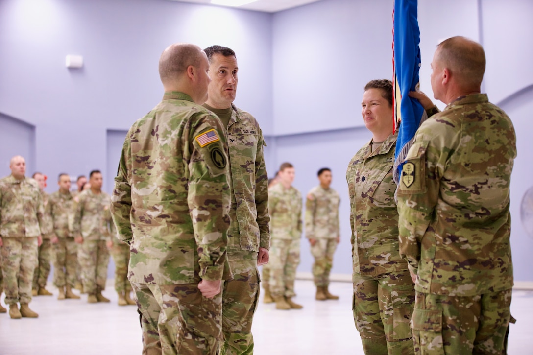 Four Soldiers stand at attention. One Soldier holds the battalion colors, a blue flag. Soldiers are seen standing in the background, slightly out of focus.