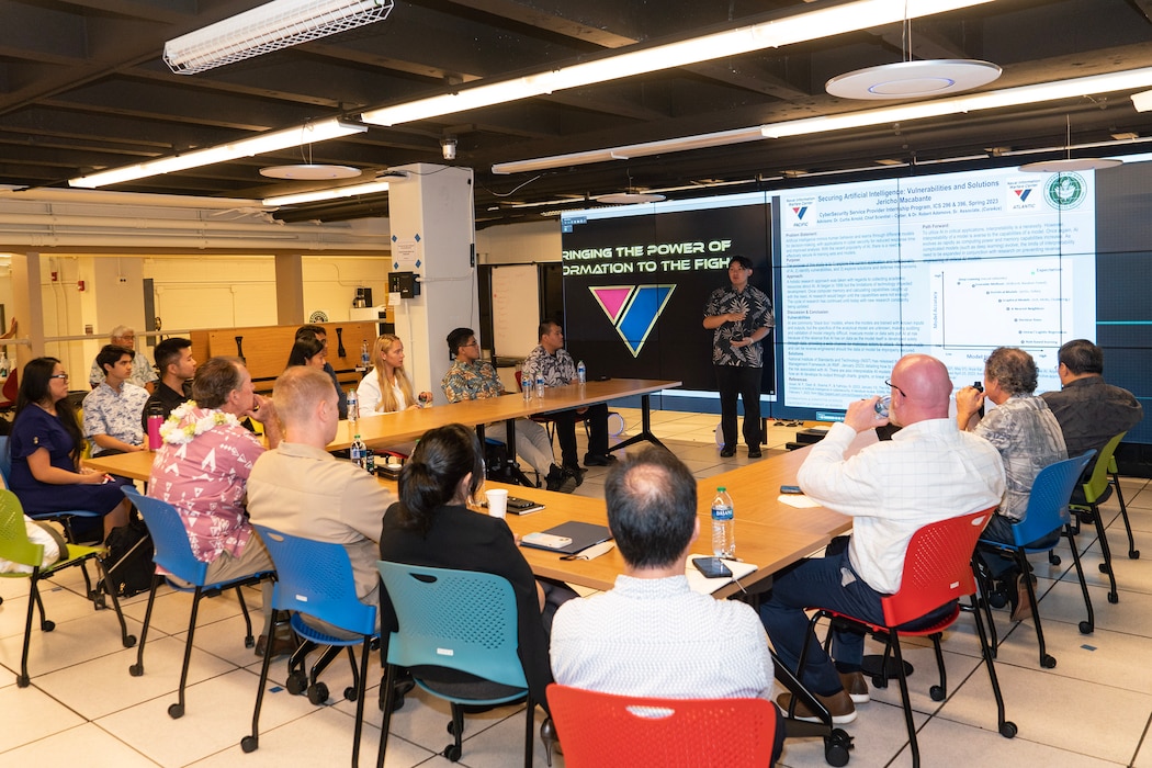 Group of people at table listening to Jerich Macabante brief in front of a presentation on a video wall